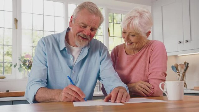 Smiling retired senior couple at home in kitchen signing legal or financial document together - shot in slow motion