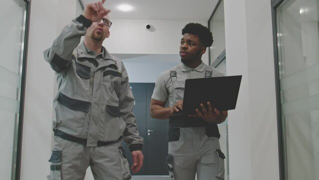 Two diverse installers in uniform walk hallway and discuss security cameras installation in modern college. Men set up CCTV cameras using computer software on laptop. Concept of surveillance system.