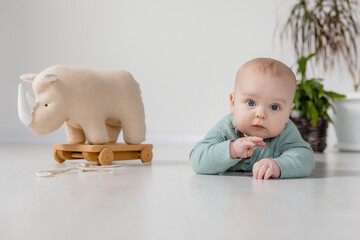 chubby baby in a green jumpsuit is lying on the floor and playing with a wooden toy on a string
