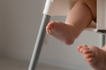 close-up of the bare feet of a baby in a green jumpsuit sitting in a white high chair for feeding. High quality photo