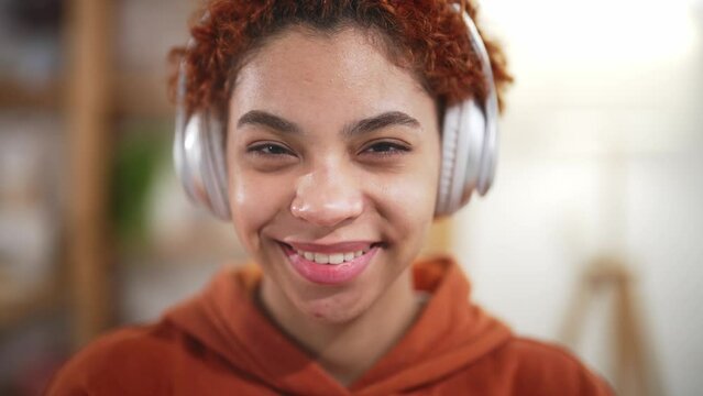 Portrait of beautiful african american young woman, sits at home on bed in headphones and listens to music. Girl listening music in headphones, sitting on sofa. Chill out and leisure concept.