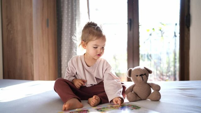 Little girl showing picture book to teddy bear while sitting on bed