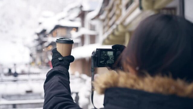 Asian woman tourist using camera taking picture of hot coffee cup during travel Ginzan onsen area in Yamagata prefecture, Japan in snow day. Attractive girl travel landmark village on winter vacation.
