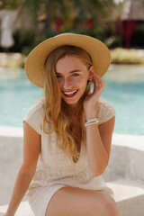 Pretty smiling blond  traveling woman in straw hat posing  near swimming pool . White summer clothes. Perfect wavy hairs.