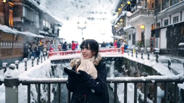 Asian woman looking snowflakes falling down during travel at Ginzan Onsen area street in Yamagata prefecture, Japan in snow day sunset. Attractive girl travel local village landmark on winter vacation