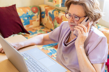 Beautiful senior old woman sitting at home with laptop and smartphone. Elderly using cellphone looking to screen. Older people learning to use new technologies on retirement. Active modern lifestyle.