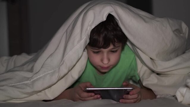 boy plays on the phone at night. a child on a bed under a blanket uses gadgets. preteen boy looks at the screen