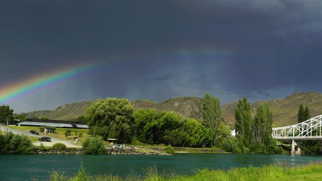 Bridge near town Alexandra in New Zealand with massive dark storm clouds and rainbow above, pan left view