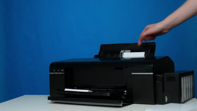 Front view of a black printer and blank sheets of a6 photo paper on a white table on a blue background, mockup. Office equipment. Copy space. Layout.