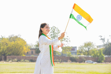 Young beautiful woman wearing traditional white dress holding indian and weaving flag while standing at park celebrating Independence day or Republic day.
