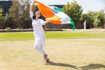 Happy young woman wearing traditional white dress running with big indian flag outdoor at park,...