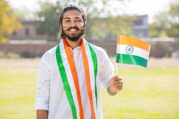happy young beard man wearing traditional white dress holding indian and weaving flag while standing at park celebrating Independence day or Republic day.