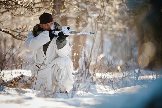 A hunter wearing  a cold-weather camouflage outfit aims a rifel while crouching in a snow-covered forest.