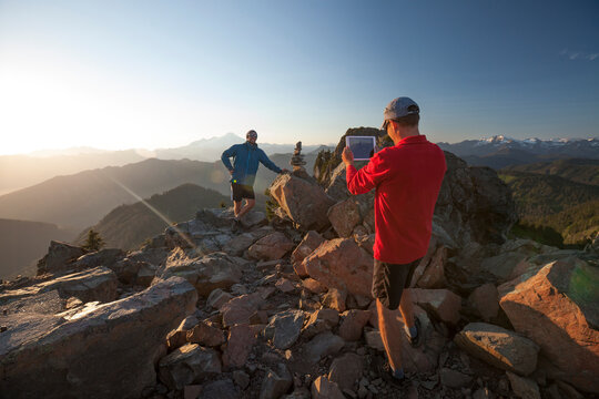 A man uses a tablet to take a picture of his friend next to a summit cairn.