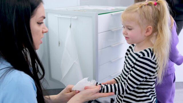 nurse doctor or teacher wipes hands with wet napkin to child talking about clean hygiene sterility in background another child with mother white furniture towels hospital school kindergarten