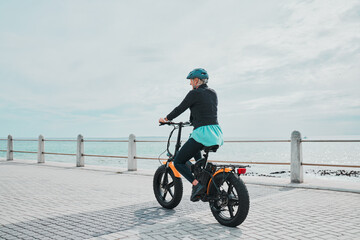 Mature woman, helmet or electrical bike by beach for future fitness, clean energy transport or sustainability travel. Old person, ebike or electric bicycle with head safety for eco friendly cycling