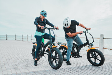 Mature couple, electrical or checking bike by beach in bonding transportation, clean energy or sustainability city travel. Ebike, electricity or eco friendly bicycle for elderly man or cycling woman