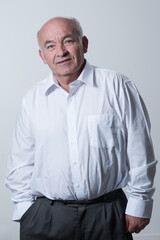 Portrait of an older man wearing a white shirt on a gray background. A healthy old man looks at a camera isolated over a gray wall. An older man smiles at the camera, a happy old man. 