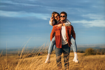 A boy is holding a girl on his back while walking through golden yellow meadow on a sunny day. They are wearing a sunglasses and smiling.