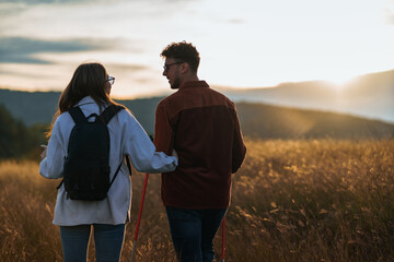A girl is holding a boy's arm during their hike through a big golden yellow meadow on a sunset, talking to each other and wearing sunglasses.