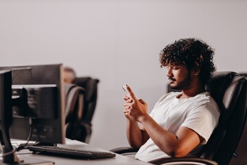 Office worker typing message on smartphone. Curly young man sitting at the computer desk in office and using his phone