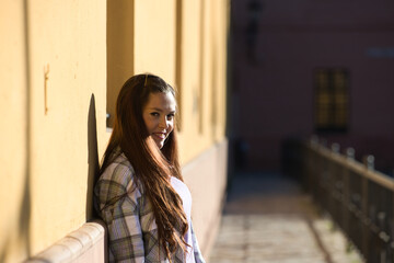 Beautiful young Spanish woman leaning against yellow wall. The woman is happy and enjoying the sunny day.