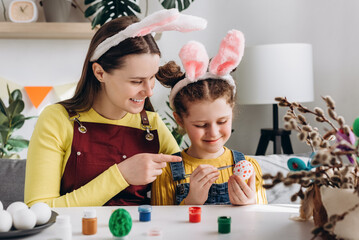 Easter celebration and traditions. Caring young mother teaching pretty little daughter child painting eggs sitting together at table, wears bunny ears. Concept of creativity and children's education