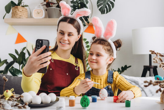 Pretty little daughter and happy young mother in fluffy bunny ears holding video call with relatives or friends using smartphone app, congratulating with Easter online, distant festive communication
