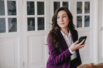 Exhausted brunette European businesswoman in violet suit sitting on leather couch holds phone looks aside with pensive face tired at home. Puzzled Hispanic woman feels fatigue. Failure, mistake.