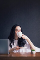 Happy casual beautiful brunette woman working on a laptop and drink coffee tea from white cup wear white t-shirt sitting on the bed in the house.
