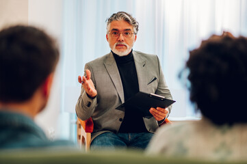 Senior man psychiatrist talking with his patients during therapy session - obrazy, fototapety, plakaty