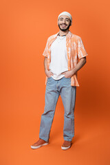 full length of happy bearded man in trendy shirt and white beanie posing with hands in pockets of blue jeans on orange background.