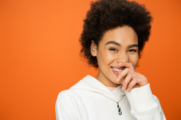 portrait of cheerful african american woman holding hand near face and looking at camera isolated on orange.