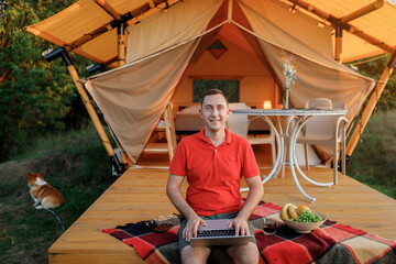 Fototapeta na wymiar Happy man freelancer programmer using a laptop on a cozy glamping tent in a summer day. Luxury camping tent for outdoor holiday and vacation. Lifestyle concept