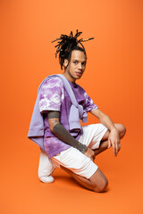 full length of multiracial man in trendy outfit sitting on haunches and looking at camera on orange...