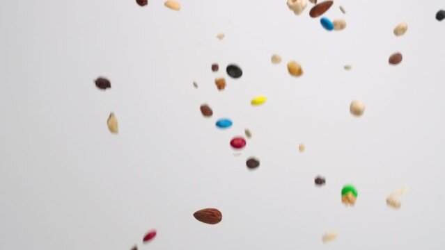 Sweet and salty trail snack mix with peanuts, pecan, almond, and cashew nuts, pretzels, m and m chocolate candies and cheerios raining down on white backdrop in slow mo