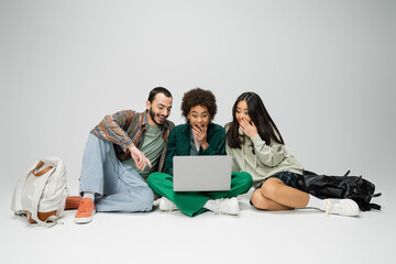 full length of amazed multicultural friends looking at laptop while sitting with crossed legs on grey background.