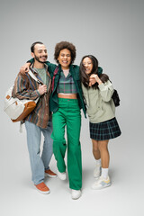 Fototapeta premium full length of joyful african american woman embracing multiethnic students with backpacks on grey background.
