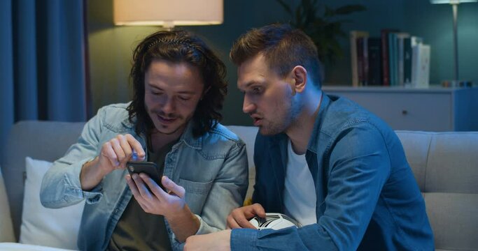 Handsome young Caucasian man showing something on smartphone to his best friend. Guys football fans putting bets at bookmakers on mobile phone in eveningt home on couch.