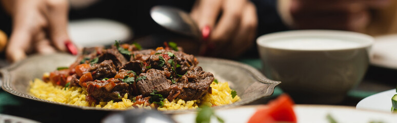 Cropped view of blurred muslim woman putting pilaf on table during ramadan dinner, banner.