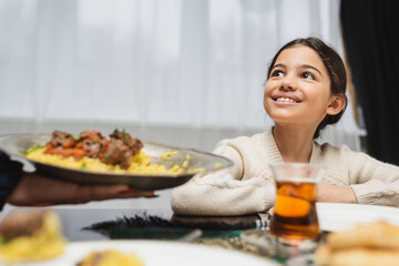 Smiling muslim girl looking at blurred mom with pilaf at home. - obrazy, fototapety, plakaty