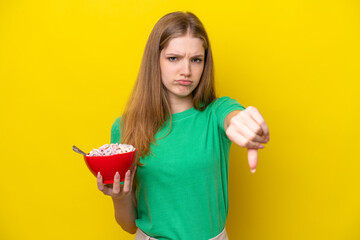 Teenager Russian girl holding bowl of cereals isolated on yellow background showing thumb down with negative expression