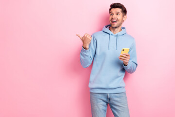 Photo of sweet impressed man wear blue hoodie browsing device looking pointing thumb empty space isolated pink color background