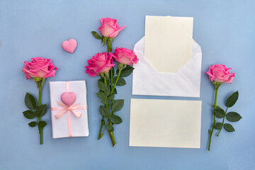 Pink flowers roses, postal envelope with paper card note with space for text, gift box with pink hearts on a blue background. Top view, flat lay