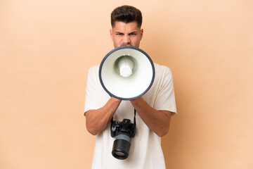 Young photographer caucasian man isolated on beige background shouting through a megaphone