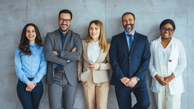 The business people standing on the gray wall background. Business team headed with boss, posing to camera over grey wall in office. Diverse businesspeople smiling at the camera