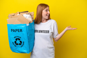 Young redhead woman holding a recycling bag full of paper to recycle isolated on yellow background with surprise expression while looking side