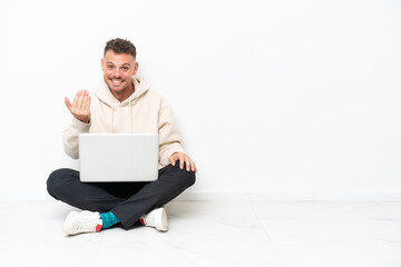 Young caucasian man with a laptop sitting on the floor isolated on white background inviting to come with hand. Happy that you came