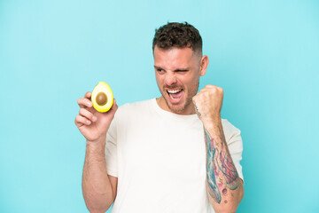 Young caucasian man holding an avocado isolated on blue background celebrating a victory