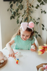 Easter. Cute little girl in bunny ears decorates Easter eggs at the table at home. Spring. Holidays.